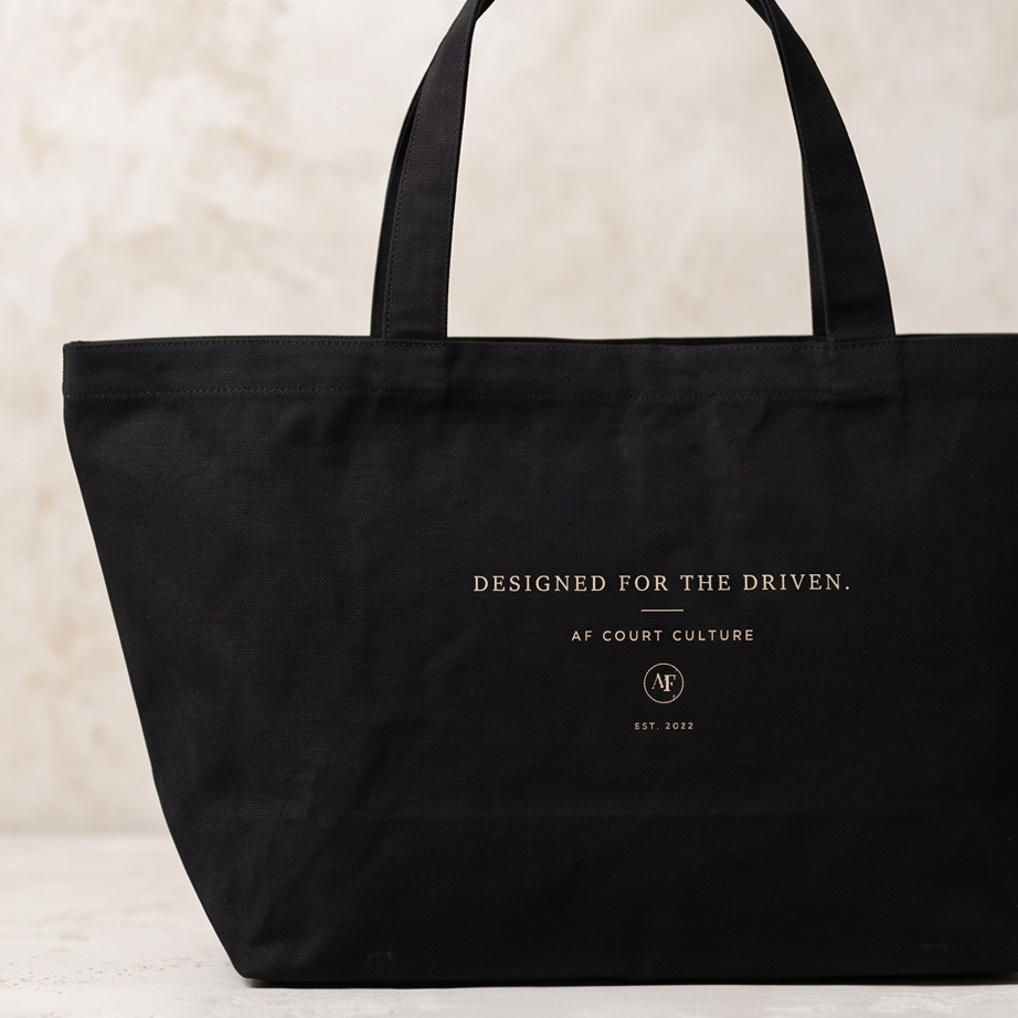 A black canvas tote bag with gold printed text reading 'DESIGNED FOR THE DRIVEN. AFCOURT CULTURE' and a circular 'AF' logo, set against a textured beige background.