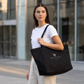 A woman wearing a white t-shirt and light beige trousers carries a black canvas tote bag with the brand name 'COURY CULTURE EST. 2026' printed in gold on the front, set against a modern glass building background.
