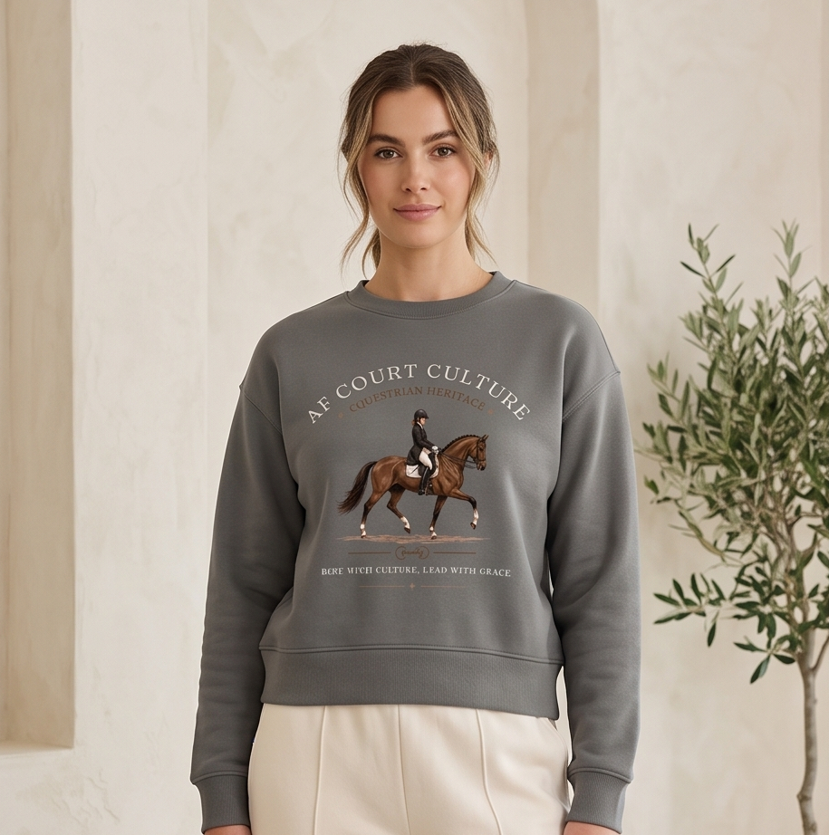 A woman wearing a gray crewneck sweatshirt with a horse-and-rider graphic and the text 'AF COURT CULTURE' and 'RIDE WITH PURPOSE LEAD WITH FACE', paired with cream pants, standing in a softly lit indoor setting with a potted plant.