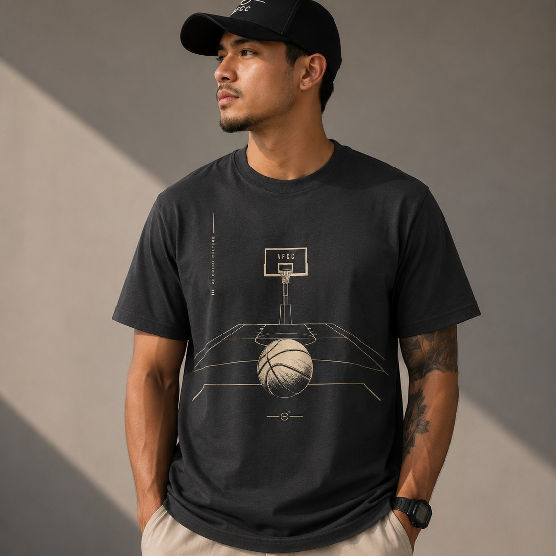 A man wearing a black crewneck t-shirt with a beige basketball court graphic, paired with beige shorts and a black baseball cap, standing against a gray wall with dramatic shadows.