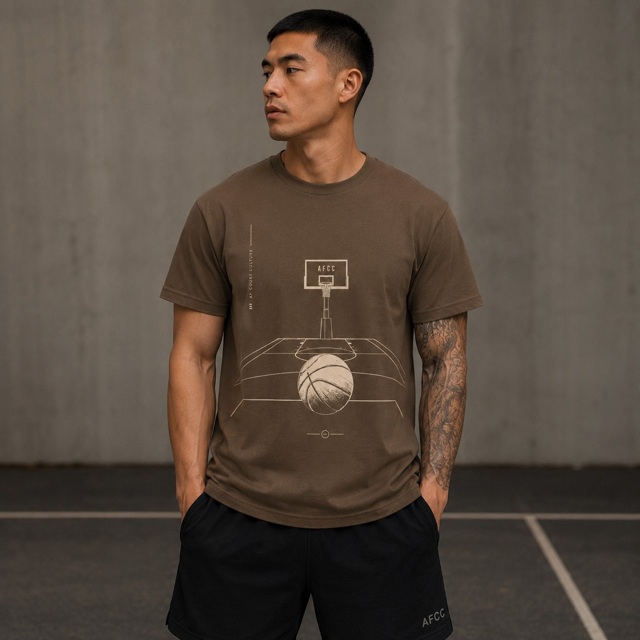 A man wearing a brown short-sleeve t-shirt with a basketball court graphic, paired with black athletic shorts, standing against a gray concrete wall.