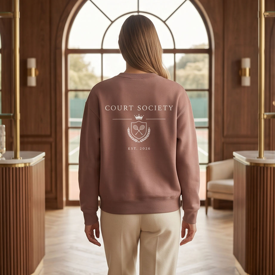 A woman viewed from behind wearing a dusty rose crewneck sweatshirt with a white 'Court Society' crest graphic on the back, paired with beige trousers, standing in a luxurious wood-paneled hall with a large arched window.