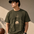 A man wearing an olive green t-shirt with a basketball court graphic, paired with beige sweatpants and a black cap, standing in a warm-lit indoor setting.