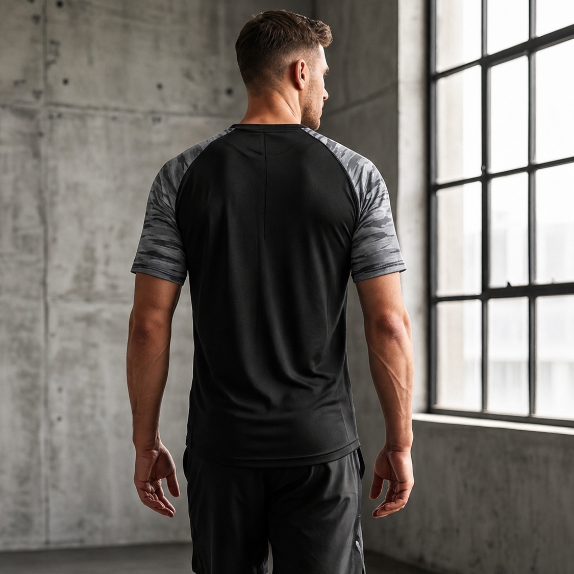 Man wearing a black athletic outfit with gray sleeves in a room with concrete walls and large windows.