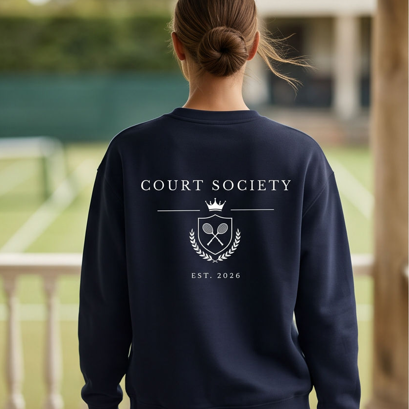 A woman viewed from behind wearing a navy blue sweatshirt with a white graphic print that reads 'COURT SOCIETY' and 'EST. 2026' on the back, standing on an outdoor patio with a tennis court blurred in the background.
