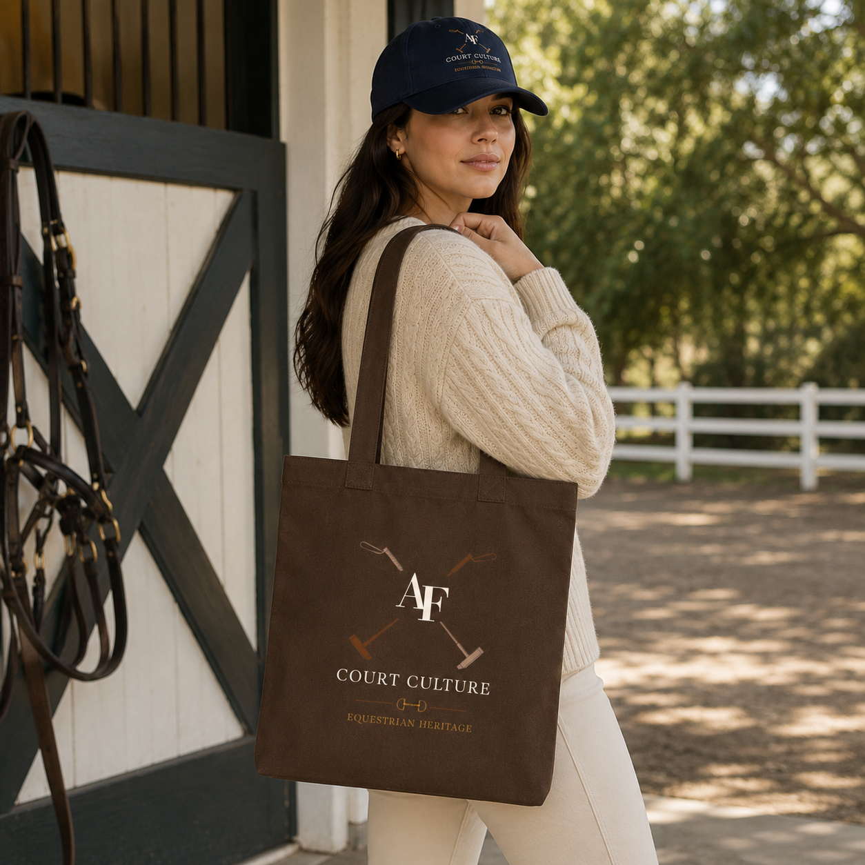 A woman in equestrian attire stands in front of a barn, carrying a dark brown canvas tote bag with a custom printed logo that reads 'AF COURT CULTURE EQUESTRIAN HERITAGE'.