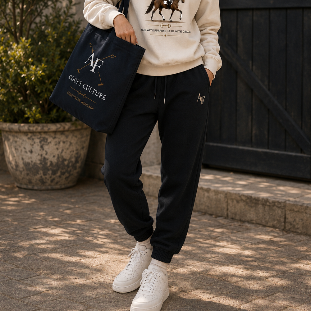 A person outdoors wearing dark navy jogger sweatpants with a small white embroidered logo, a cream sweatshirt, white sneakers, and carrying a navy tote bag with white and gold lettering.