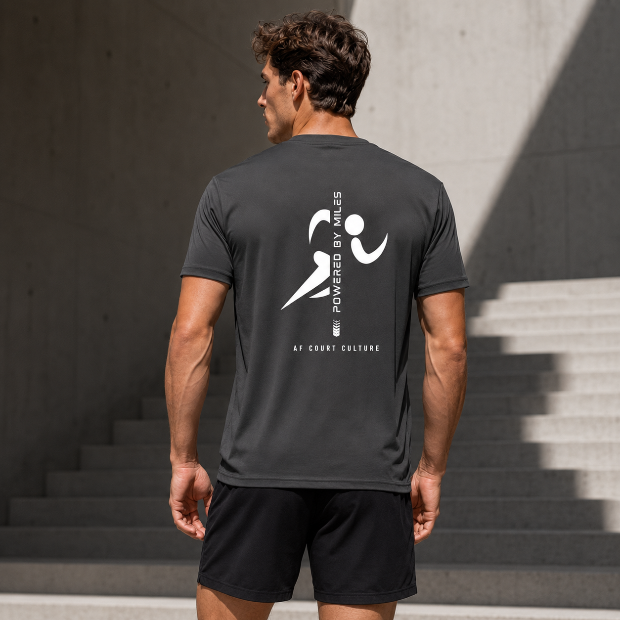 Man wearing a dark gray t-shirt with a logo and text on the back, standing outdoors.