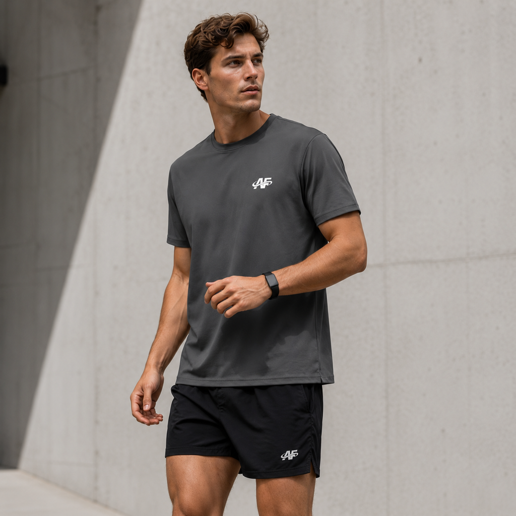 Man wearing a dark gray t-shirt and black shorts with a logo, standing against a concrete wall.